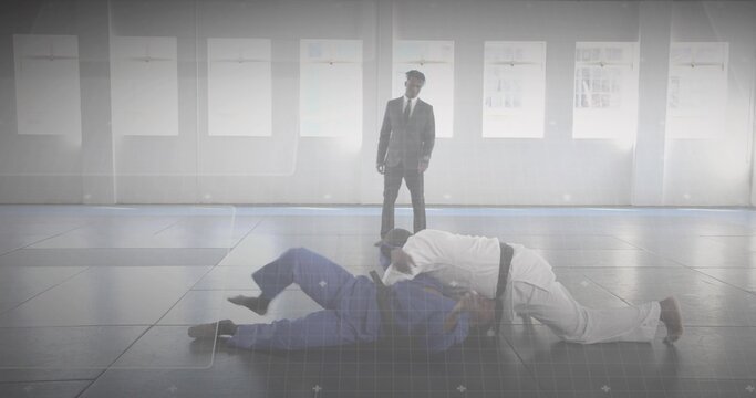 Grappling artists in gis on tatami mats near windows, with coach in suit watching