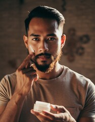 Handsome young Asian man with beard applying moisturizing facial cream to cheek, daily male grooming and skincare routine concept