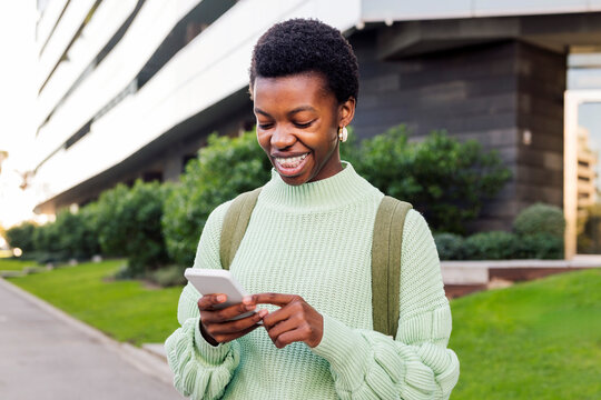 young african american woman with short hair smiling happy while looking her cell phone, concept of technology of communication and modern lifestyle, copyspace for text
