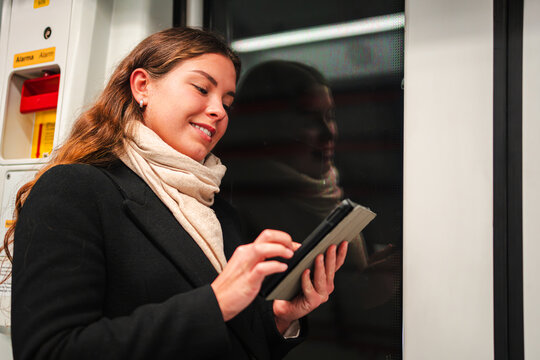 Engaged female passenger happily reading on a tablet device while commuting on public transport. She is immersed in her e-reader, showcasing a moment of concentration during her journey on the subway.