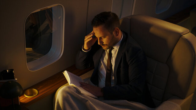 Stressed businessman in a suit having a painful headache, reading a book while traveling by private jet at night, highlighting the demands of business travel and professional burnout