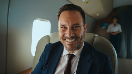 Confident businessman smiles at camera while relaxing in a luxurious private jet cabin with flight...