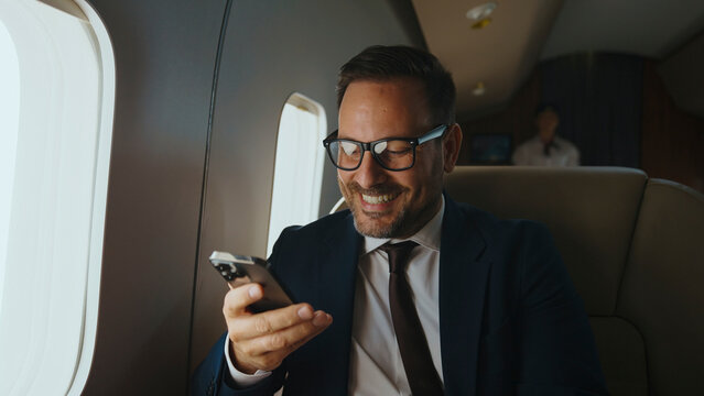 Businessman smiling happily while watching content on his mobile phone, traveling in a private jet cabin, reflecting modern business travel, luxury, and connectivity - Powered by Adobe