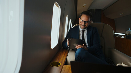 Businessman in suit traveling on a private jet, using a digital tablet for work, embodying concepts of luxury business travel, success, and productivity in a comfortable executive environment © Videophilia
