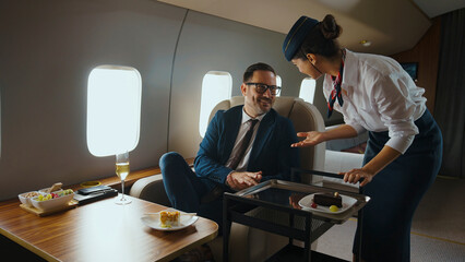 Flight attendant providing excellent service, offering a sweet dessert from a serving cart to a smiling businessman enjoying luxury travel and comfort on a private jet cabin © Videophilia
