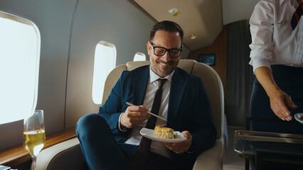 Smiling businessman in a suit enjoying a sushi meal with chopsticks while a flight attendant serves him on a luxurious private jet, embodying first-class travel and exclusive service © Videophilia