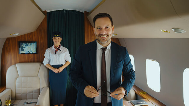 Businessman holding glasses and smiling at camera with a flight attendant standing in a luxurious private jet cabin, ready for a comfortable and exclusive business travel experience - Powered by Adobe