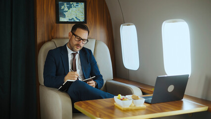 Businessman in a suit and glasses writing in a notebook while traveling on a private jet, enjoying refreshment and working with a laptop during an executive trip © Videophilia