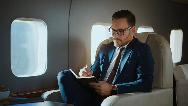 Businessman in a suit and glasses writing in a notebook, concentrating on work while flying in a luxurious private jet, embodying success and exclusive travel