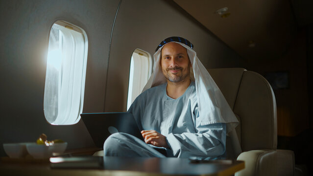 Arab businessman in traditional attire smiling while working on a laptop inside a luxurious private jet cabin, conveying wealth, global business, technology and executive travel