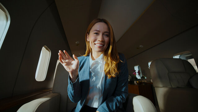 Businesswoman smiling and waving during a video call aboard a modern private jet, working remotely while traveling in luxurious, comfortable executive cabin surroundings