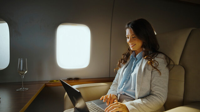 Smiling businesswoman typing on laptop in private jet cabin, blending luxury travel and professional productivity—confident executive working remotely with elegance and focus