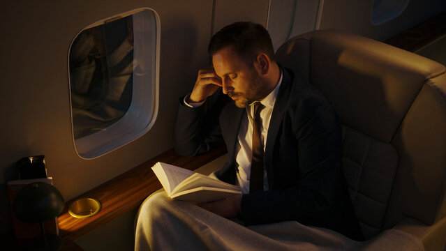 Businessman in a suit sleeping while holding a book under a blanket in a luxurious private jet cabin during a long overnight flight, finding rest during travel