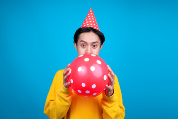 Joyful Young Man in Festive Hat Enthusiastically Inflates Polka Dot Balloon for Vibrant Celebration © Gatot