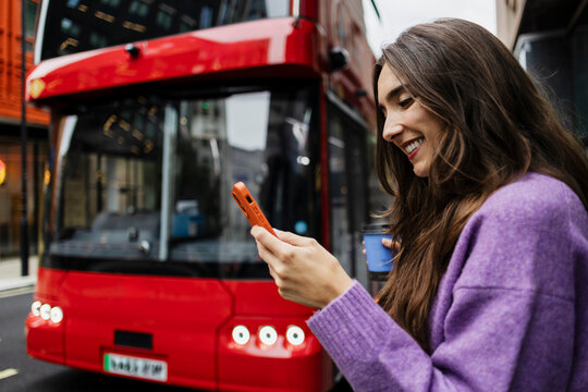 Smiling woman holding smartphone and coffee by red bus in London