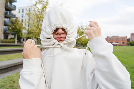 Teenager in white hoodie covering face with strings in urban park