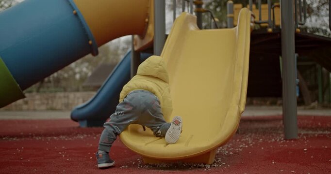 A toddler climbs up a yellow slide at a playground. The child is wearing a yellow jacket and grey pants.