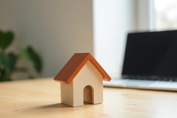 A small wooden house figurine sits on a light wood desk, with a laptop and green plant softly blurred in the background  symbolizing home, work, or real estate concepts.