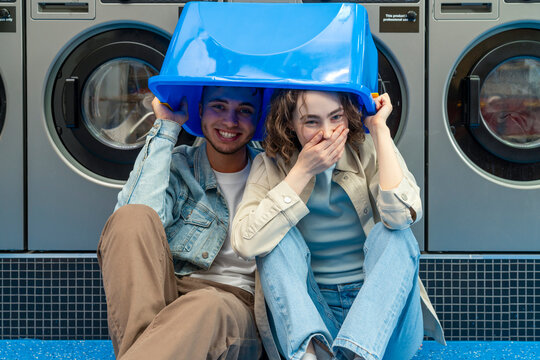 Playful young couple with blue basket over head sitting in front of washing machines at laundromat