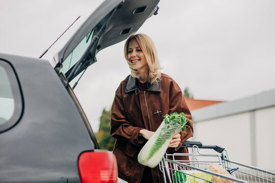 Smiling woman loading groceries into car trunk in supermarket parking