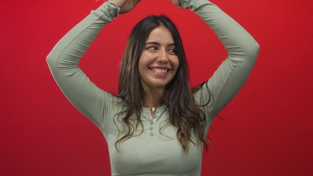 Hispanic woman in light green top smiling while making roof gesture with hands overhead against red studio wall; playfulness.