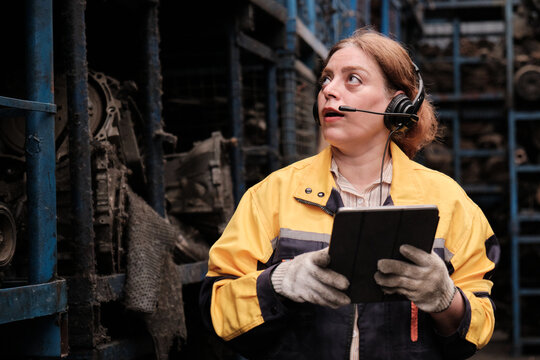 One Hispanic mature female inspector remotely works with headset and tablet, checks auto spare parts stock in warehouse stack, operator jobs in manufacturing factory, and industrial worker occupation.