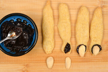 An overhead shot showing the process of decorating baked witch finger cookies by attaching almond nails with dark plum jam, next to a bowl of jam