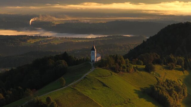 Aerial parallax of the Church of St. Primus and Felician on Jamnik Hill, Slovenia, with the Sava River blanketed in fog and the snow-capped Julian Alps glowing in the soft golden light