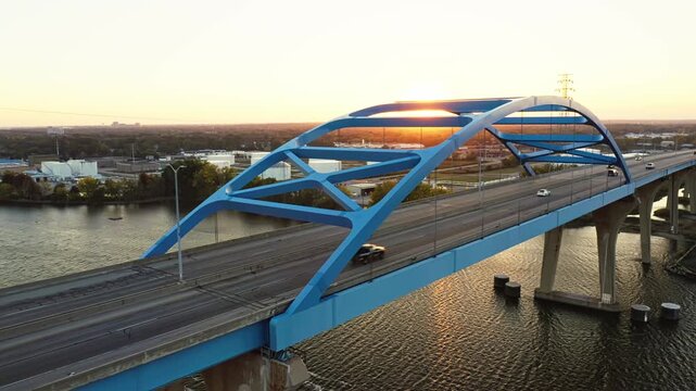 The Leo Frigo Memorial Bridge glows in the warm hues of sunset as cars cross its blue steel frame over the Fox River, reflecting golden light across the calm rippling water.