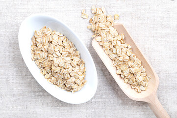Rolled oats, thick-rolled and old-fashioned oats, in white bowl and grain scoop, on linen. Dehusked and steamed oat groats, rolled into flakes under heavy rollers, stabilized through lightly toasting.