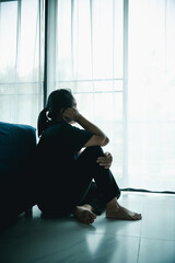 A lonely woman sits on the floor by window with hand on head in dark room. Concept of depression, sadness, anxiety, stress, mental health problem, loneliness and emotional struggle in life.
