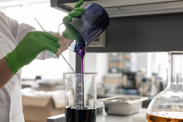 A laboratory technician in green nitrile gloves pour a dark purple liquid from one beaker into another. A сraft production process of organic cosmetics, extraction process, mixing ingredients in a lab