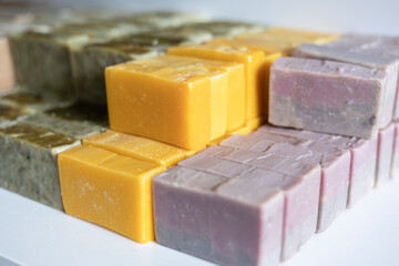 Various bars of handmade, cold-process soap are curing on a shelf. These organic cosmetics feature different natural ingredients, resulting in beige, green, yellow, and purple colors.