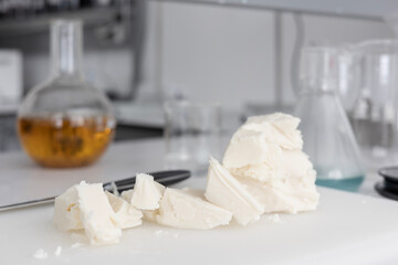 Raw shea butter, cut into chunks, sits on a white cutting board in a laboratory. This ingredient is for the craft production of organic cosmetics, with blurred glassware and extracts in the background