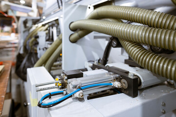 A close-up of an automated edge banding machine in a furniture factory. Corrugated plastic hoses for dust extraction are connected to the machine, which glues and cuts edges on MDF and chipboard.