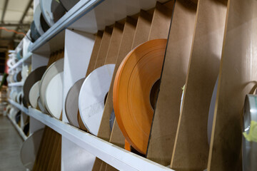 A close-up of shelves in a furniture factory warehouse, stocked with many large spools of plastic edge banding tape in various colors and textures for manufacturing.
