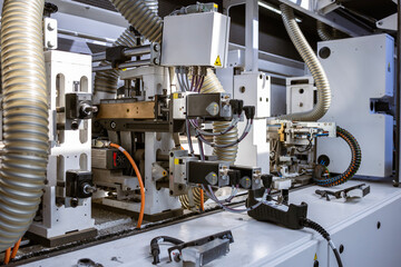 A close-up view of the internal mechanics of an automated edge banding machine. It shows various motors, wiring, and corrugated hoses for dust collection in a furniture factory.