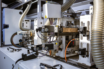 A close-up view of the internal mechanics of an automated edge banding machine. It shows various motors, wiring, and corrugated hoses for dust collection in a furniture factory.