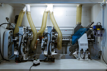 A wide view of the internal mechanics of an automated edge banding machine in a furniture factory. It shows various motors, rollers, and yellow corrugated hoses for dust collection.