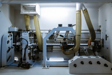A wide view of the internal mechanics of a automatic edge banding machine in a furniture factory. It shows the rollers, motors, and yellow corrugated hoses for dust collection.