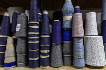 A close-up view of yarn cones on a shelf in a knitwear production warehouse. The shelf is stocked with bobbins of various blue, grey, and purple threads, the raw material for knitting.