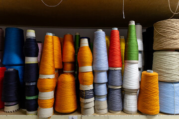 A close-up view of large yarn cones stacked on a shelf in a knitwear factory. The colorful bobbins of white, brown, blue, and orange thread are raw materials for manufacturing.