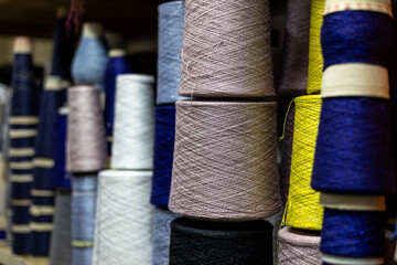 A close-up, selective focus view of yarn cones in a knitwear factory. Large bobbins of mauve, blue, yellow, and white thread are stacked on a shelf, ready for manufacturing.