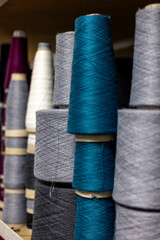 A close-up, selective focus view of yarn cones in a knitwear factory. Large bobbins of teal, grey, and white thread are stacked on a shelf, ready for manufacturing.