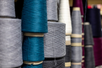 A close-up, selective focus view of yarn cones in a knitwear factory. Large bobbins of teal, grey, and white thread are stacked on a shelf, ready for manufacturing.