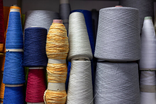 A close-up, colorful arrangement of yarn cones in a knitwear factory. Large bobbins of blue, yellow, grey, and red thread are stacked on a shelf, showing the raw materials for manufacturing.