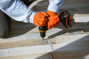 A close-up of a worker's hands in orange gloves using a cordless electric drill to drive a screw into a new wooden deck. This DIY construction project involves assembling planks outdoors.