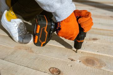 A close-up of a worker's hands in orange gloves using a cordless electric drill to drive a screw...