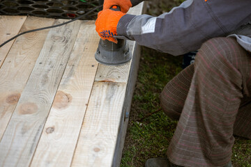 A worker in an orange glove uses an electric orbital sander to smooth new, light-colored wooden planks. This DIY woodworking project involves preparing a deck or floor surface outdoors.