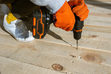 A close-up of a worker's hands in orange gloves using a cordless electric drill to drive a screw into a new wooden deck. This DIY construction project involves assembling planks outdoors.